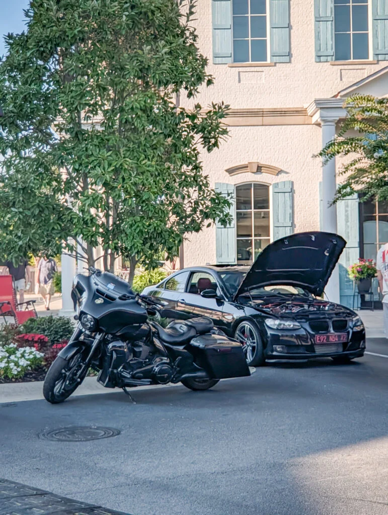 Black BMW coupe with open hood and black touring motorcycle parked on street near white house with blue shutters