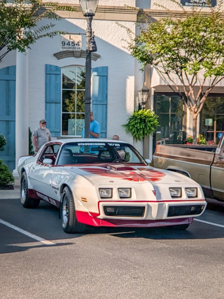 White and red Pontiac Trans Am with hood decal parked in front of a building with blue shutters and trees
