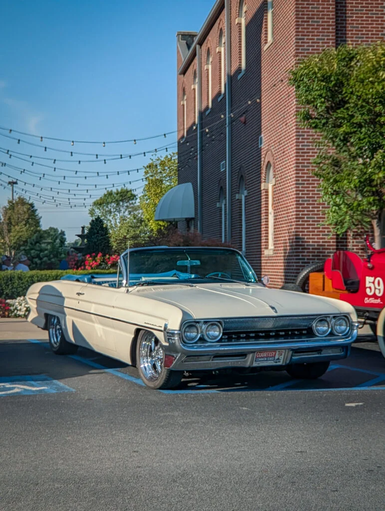 White Oldsmobile Starfire Convertible parked beside a brick building with outdoor string lights overhead