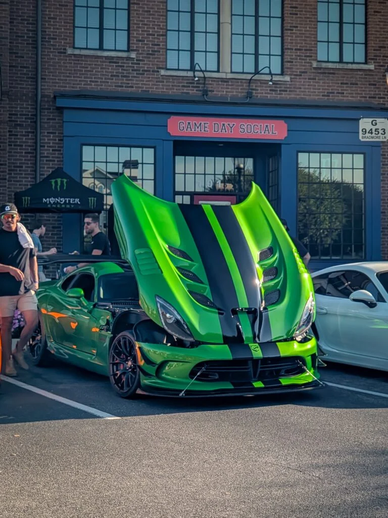 Green Dodge Viper with black racing stripes and open hood parked in front of a brick building with people nearby