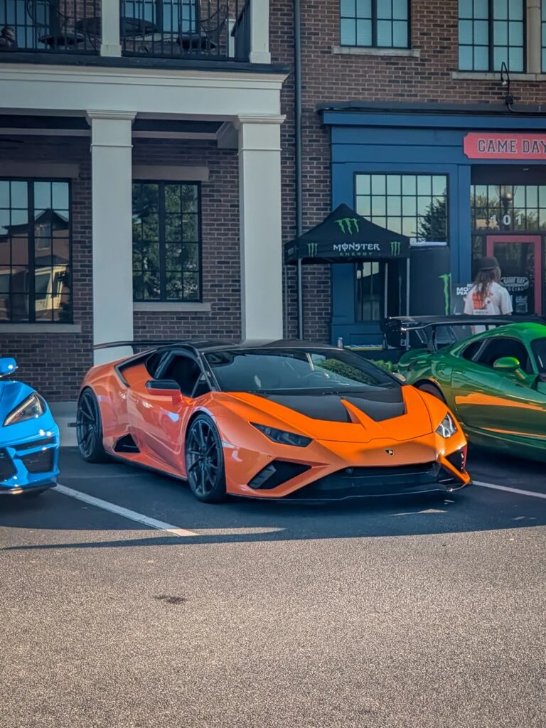 Orange Lamborghini Huracán with black hood and roof parked beside a blue and green car in front of a brick building