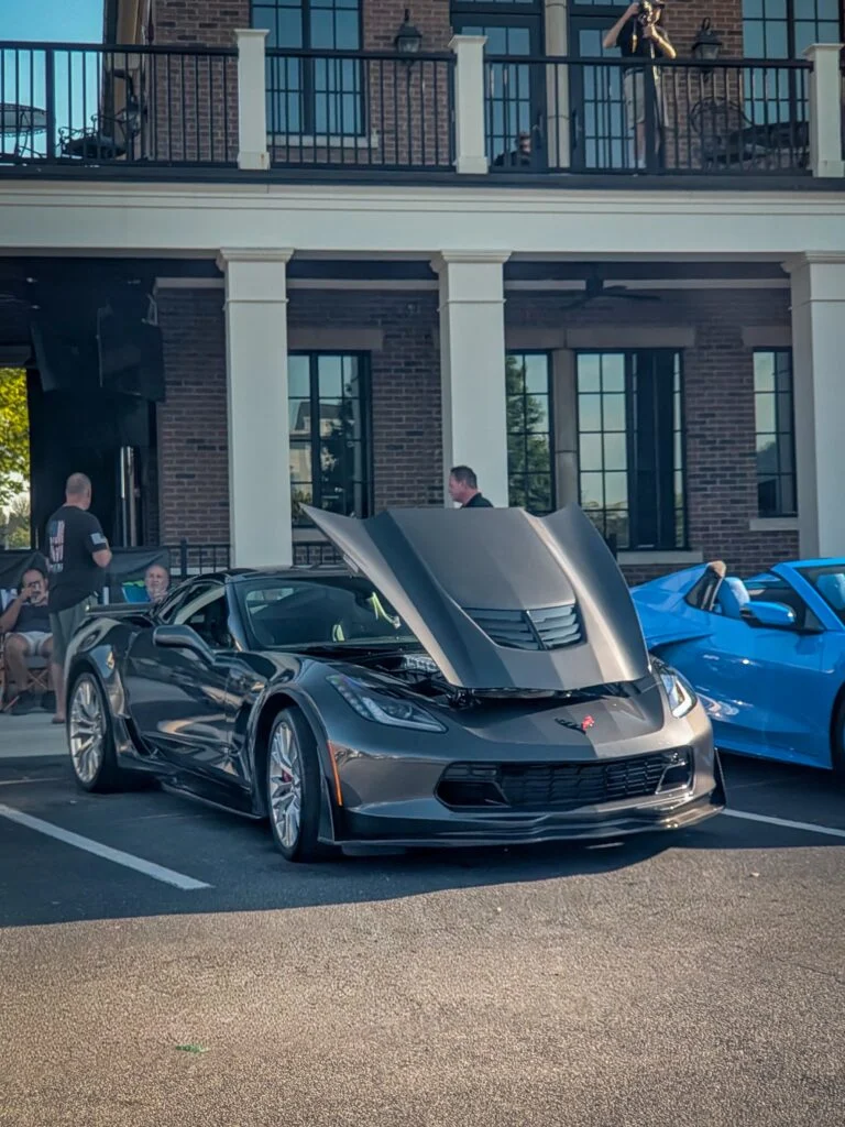 Gray Chevrolet Corvette with hood open at an outdoor event parking lot, people nearby and building with windows in background