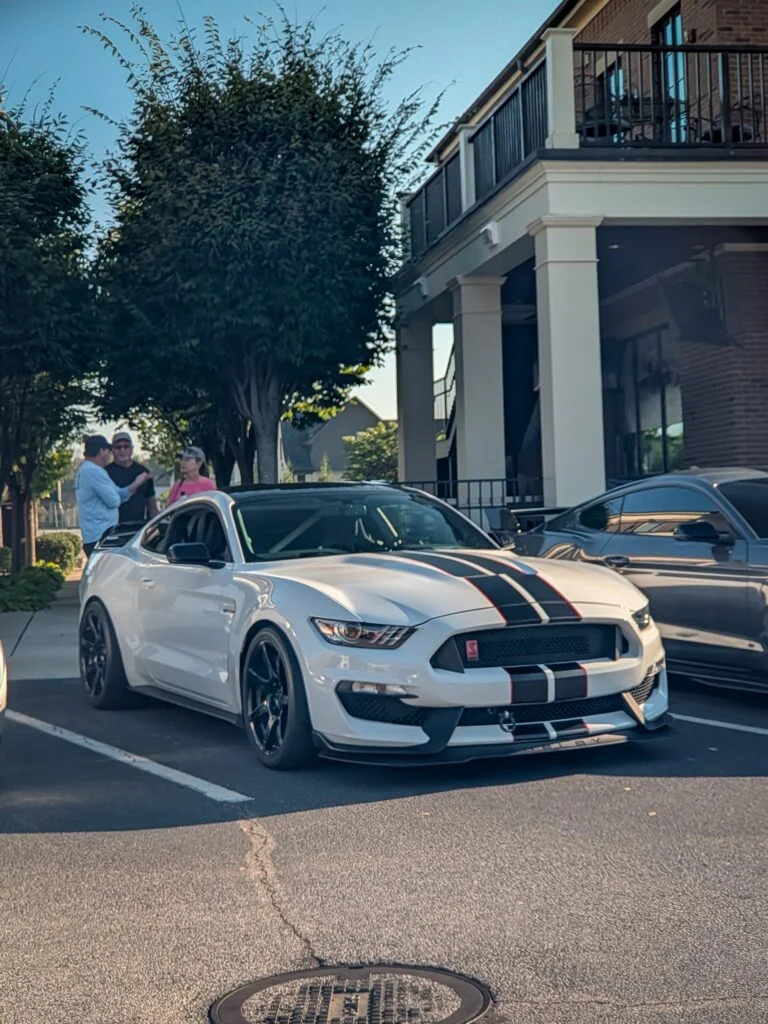White Ford Mustang Shelby GT350R with black and red stripes parked on street, three people chatting nearby under trees