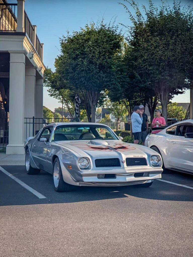 Silver Pontiac Firebird Trans Am parked in a lot with people and trees in the background