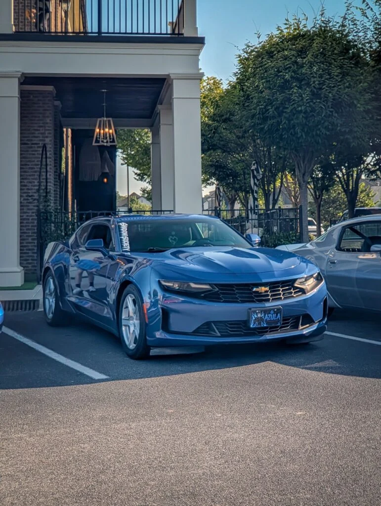 Blue Chevrolet Camaro parked next to a white classic car in an outdoor setting with trees and building columns