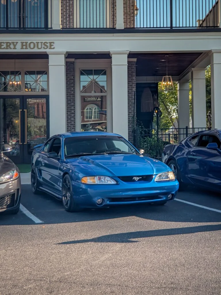 Blue Ford Mustang parked outside a brick and white-column building under daylight.