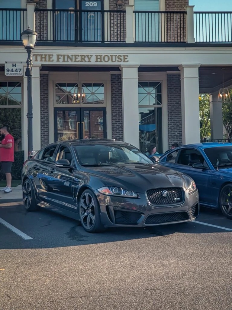 Dark gray Jaguar XFR-S sedan parked in front of The Finery House with other vehicles and people nearby
