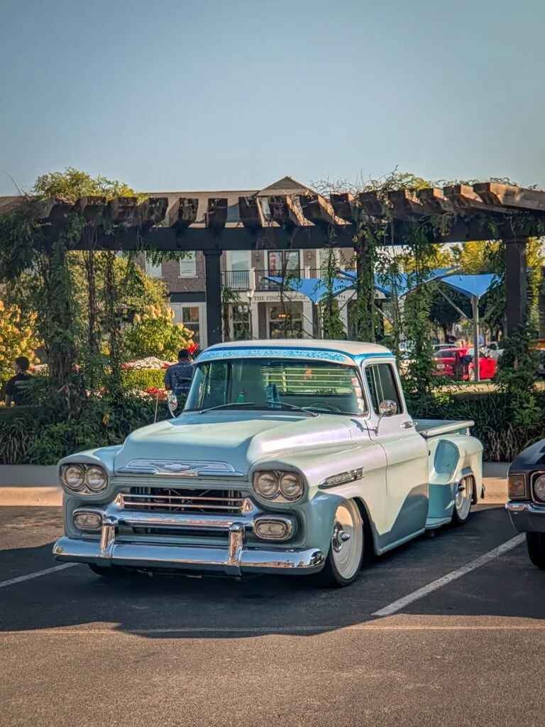 Light blue Chevrolet Apache truck parked under a wooden pergola at an outdoor car event.