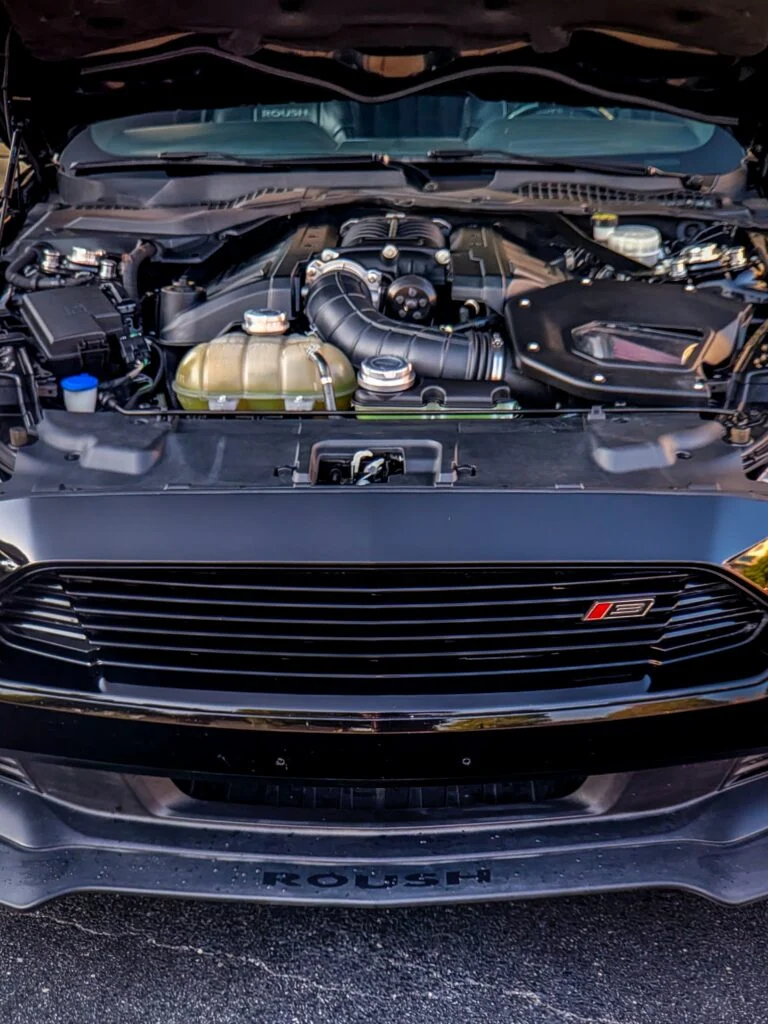 Engine bay of a black Roush Ford Mustang with a ROUSH badge on the front grille visible, hood open outdoors.