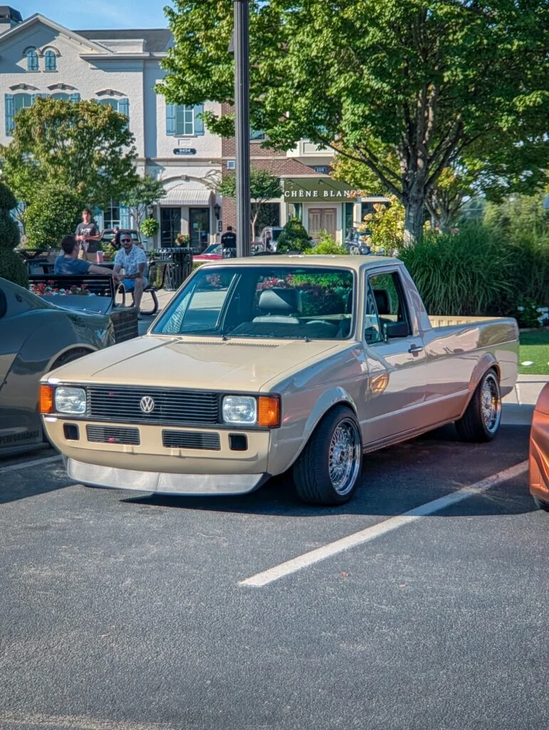 Cream-colored vintage Volkswagen Rabbit pickup truck parked in a lot with people and buildings in the background