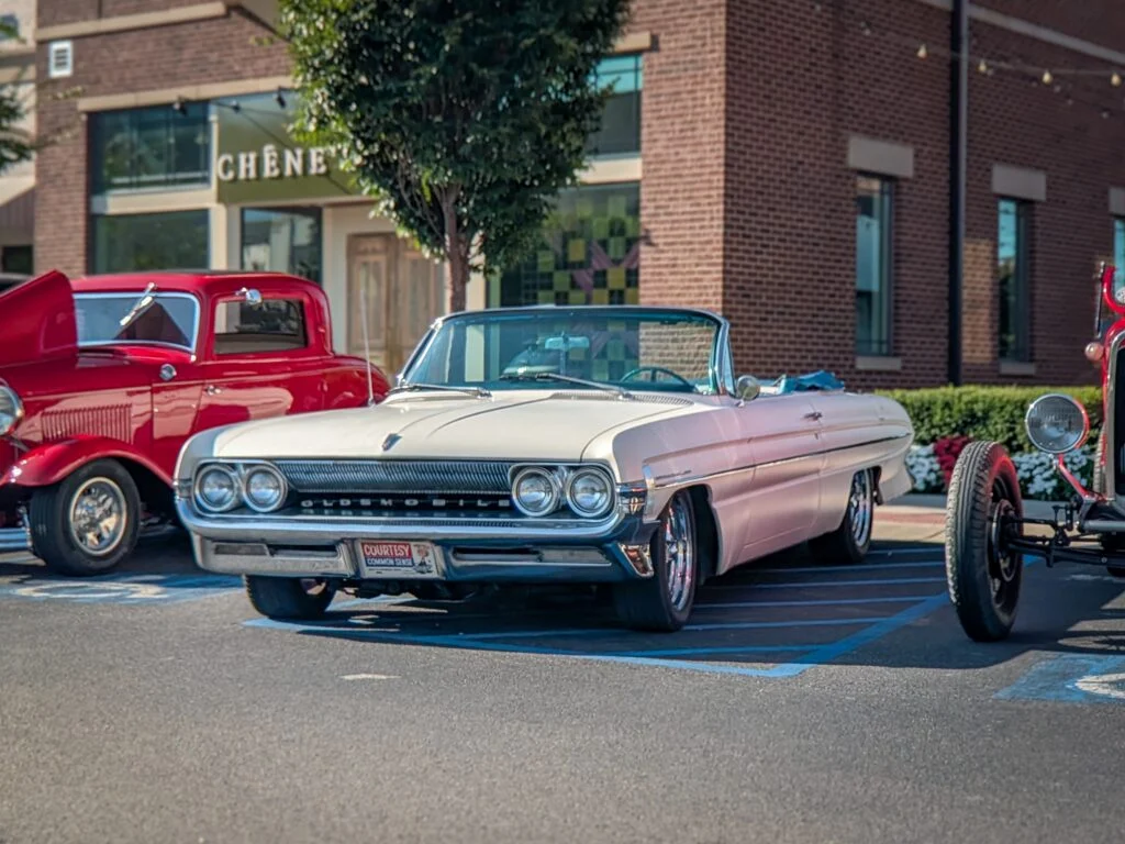 White Oldsmobile Starfire Convertible parked in front of a brick building with a red hot rod and a red classic truck nearby