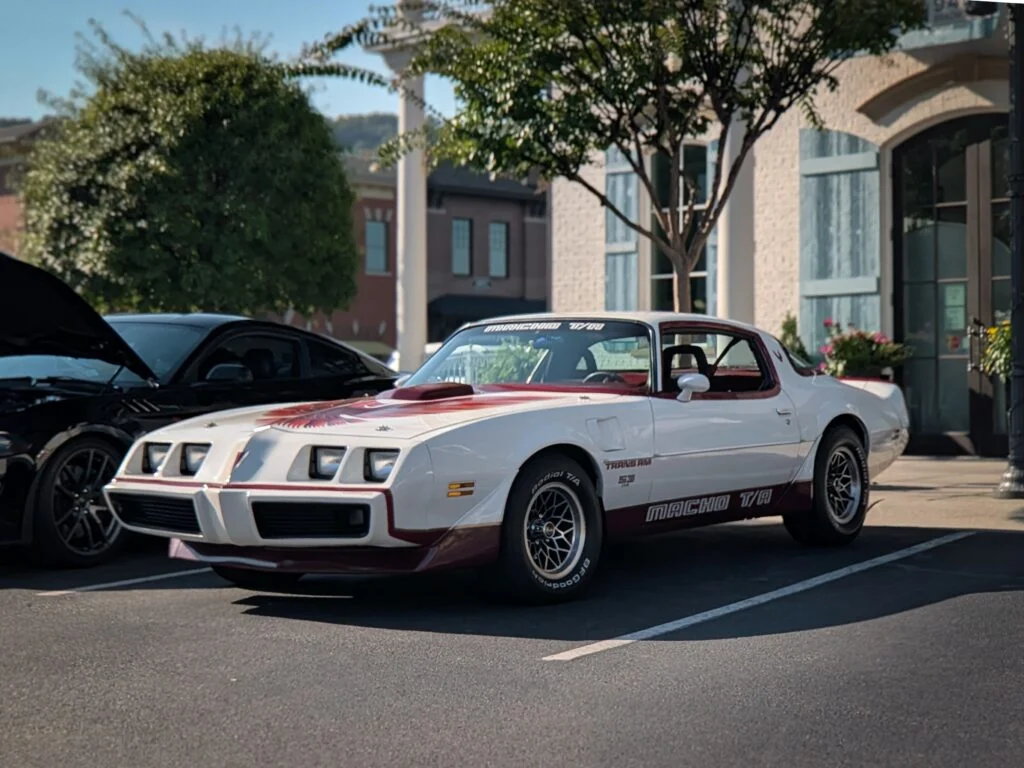 White Pontiac Trans Am Macho T/A with decals parked in front of buildings and trees at a car event