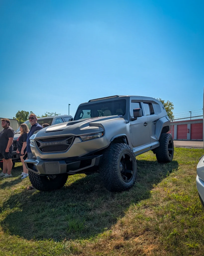 Silver Rezvani Tank parked outdoors with a group of people standing beside it under a clear blue sky