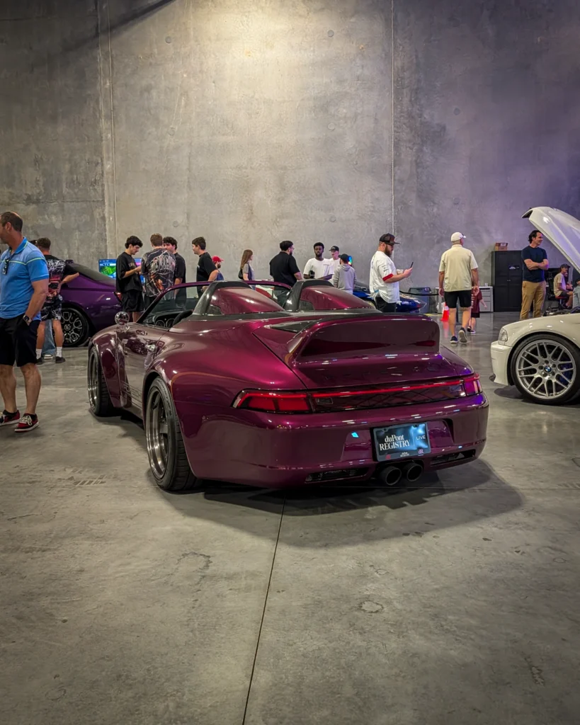 Maroon Porsche Speedster and white BMW sedan in an indoor car event with people standing and walking nearby
