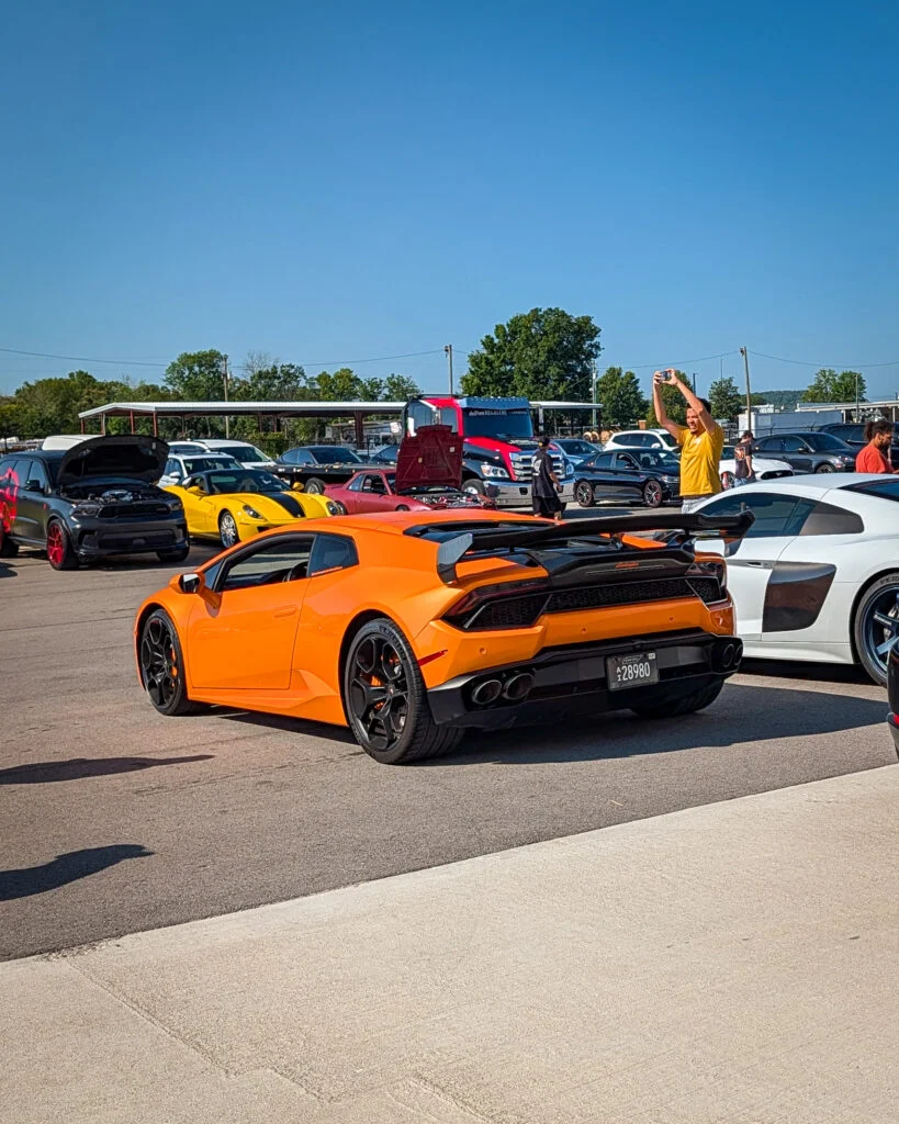 Orange Lamborghini Huracán and white Audi R8 parked at a car event with people and other vehicles in the background