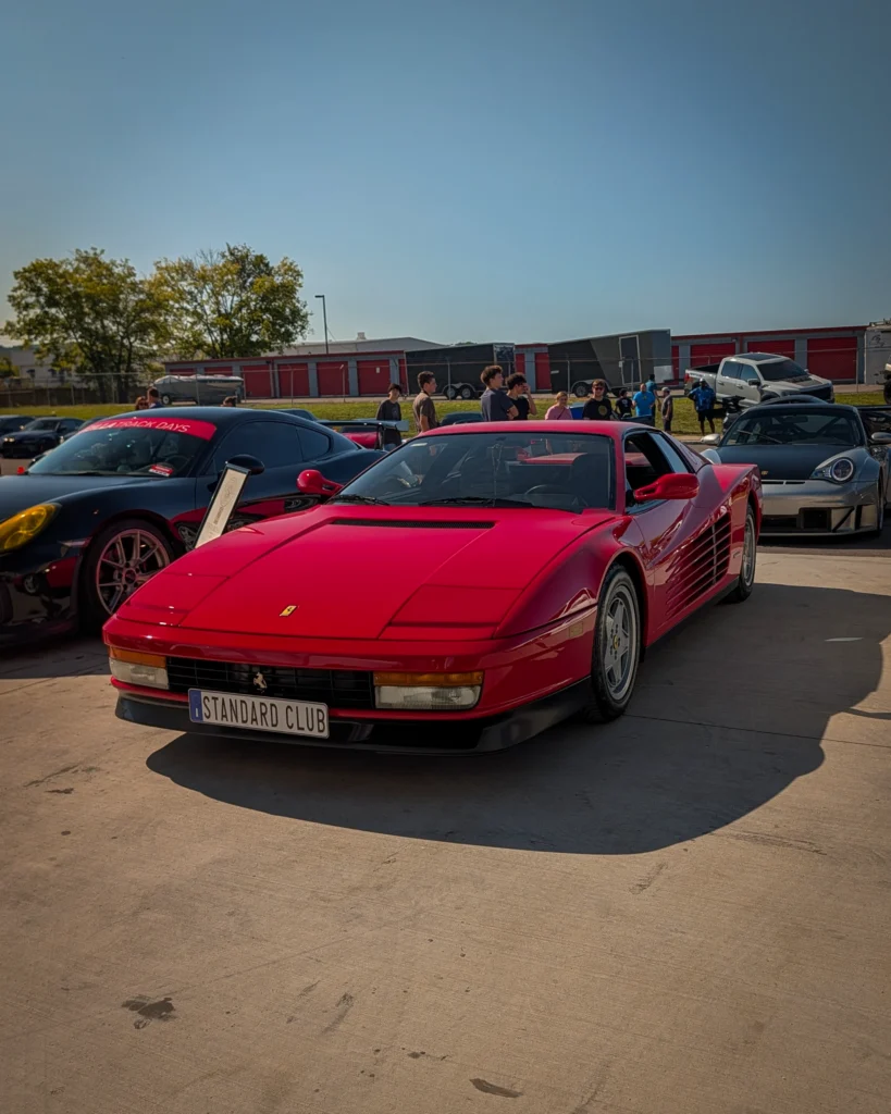 Red Ferrari Testarossa with black interior parked on concrete under clear blue sky at car event with people and other cars