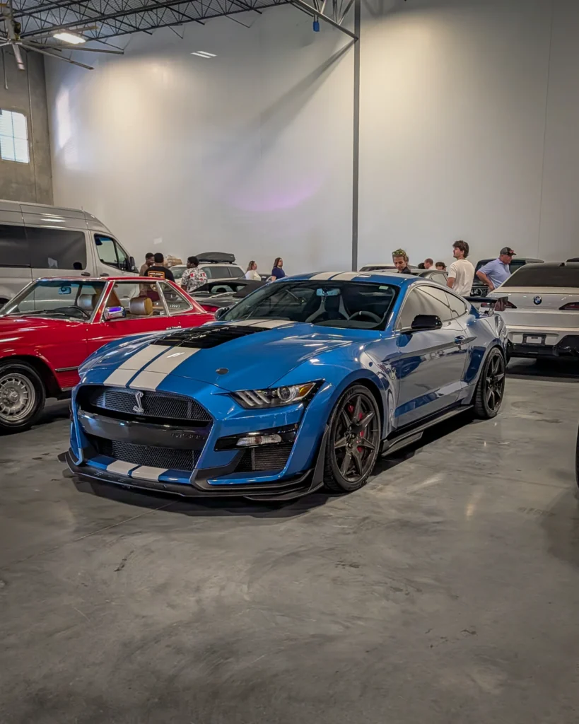 Blue Ford Mustang Shelby GT500 with white stripes parked inside a building with people and other cars nearby