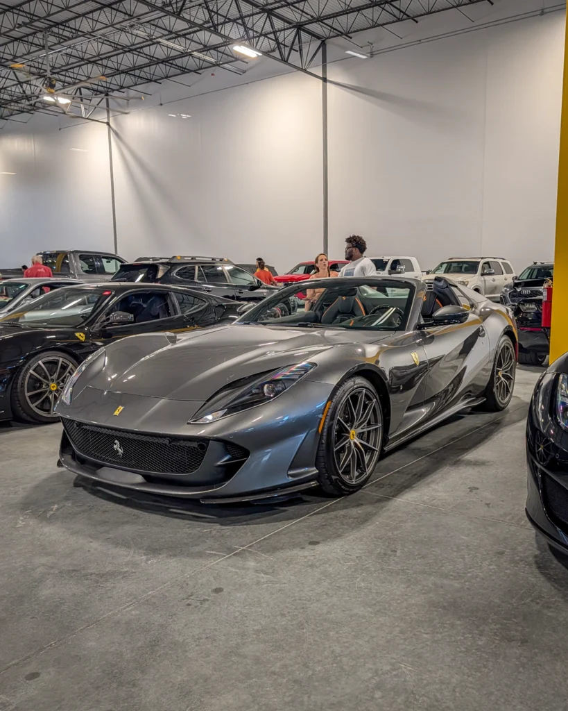 Gray Ferrari 812 GTS parked indoors at a car event with people and other vehicles in the background