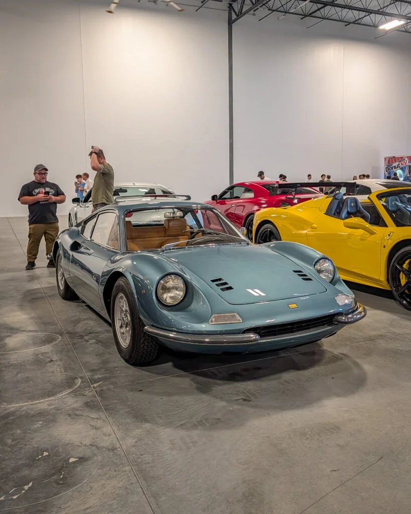 Blue Ferrari Dino coupe in an indoor car show with other colorful sports cars and people in background