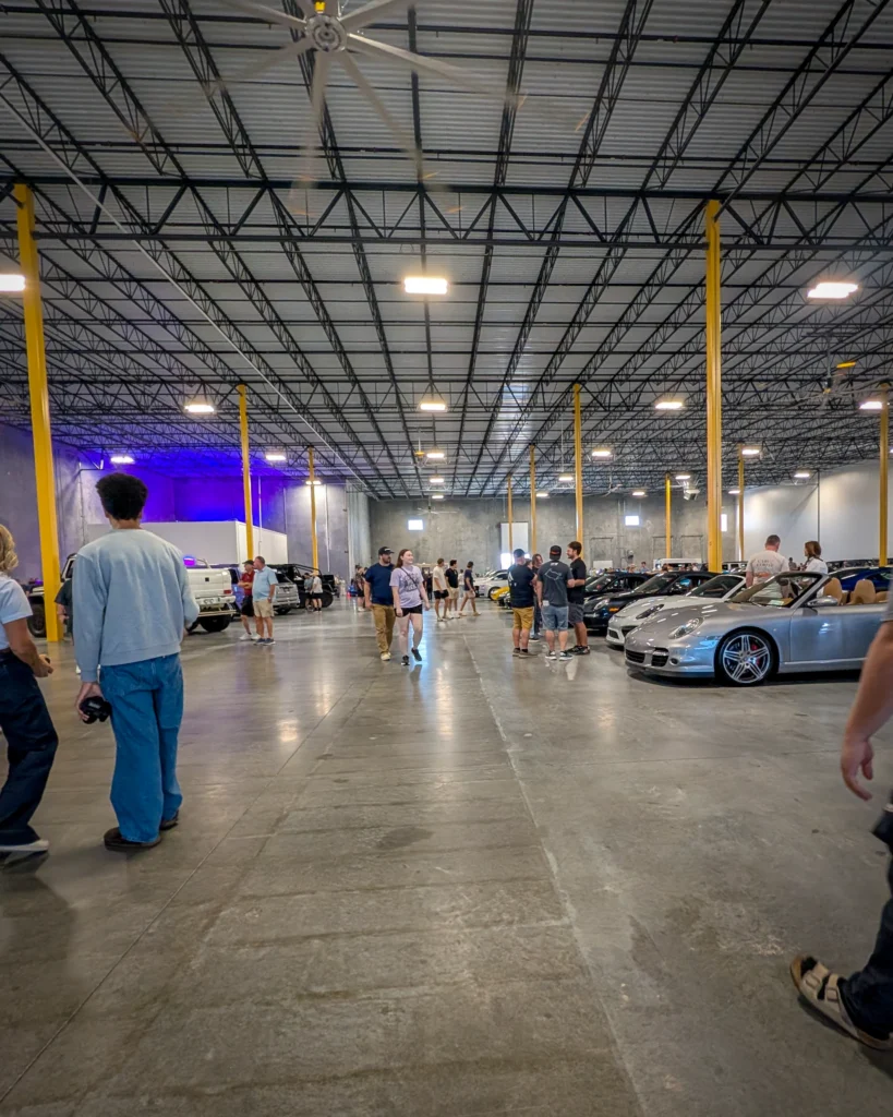 People walking and standing among parked cars in a large warehouse with yellow support poles and a concrete floor.