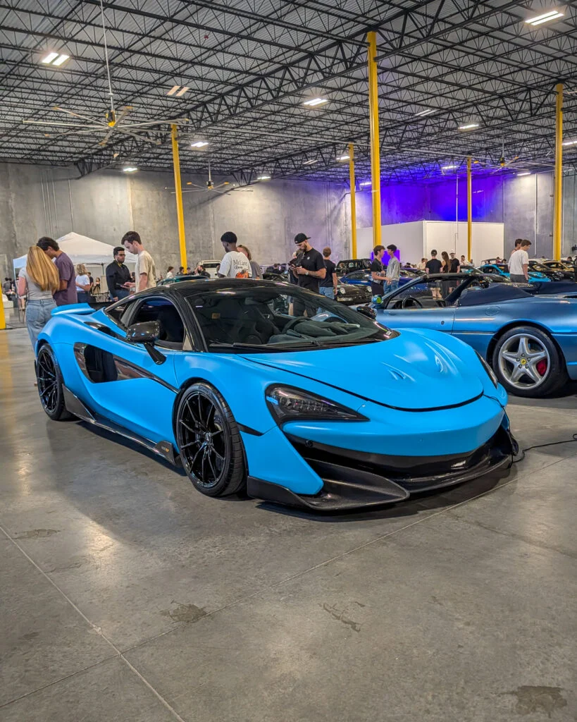 Blue McLaren 600LT with black roof parked indoors surrounded by people and other vehicles at a car event