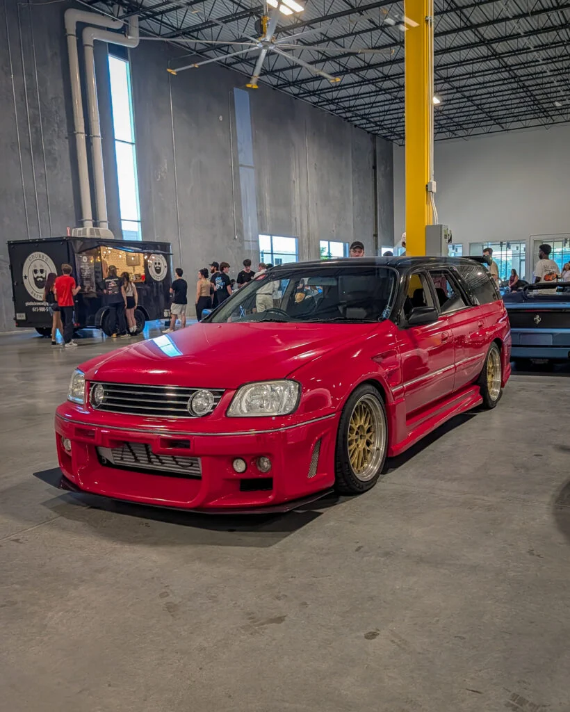 Red Nissan Stagea with black roof parked indoors at a car event with people and a food truck in the background