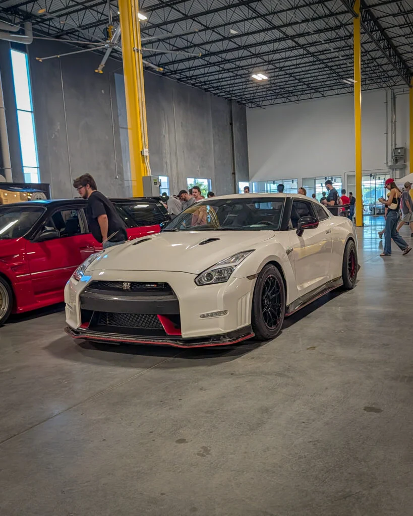 White Nissan GT-R Nismo parked indoors at a car event with people walking around and a red car in the background