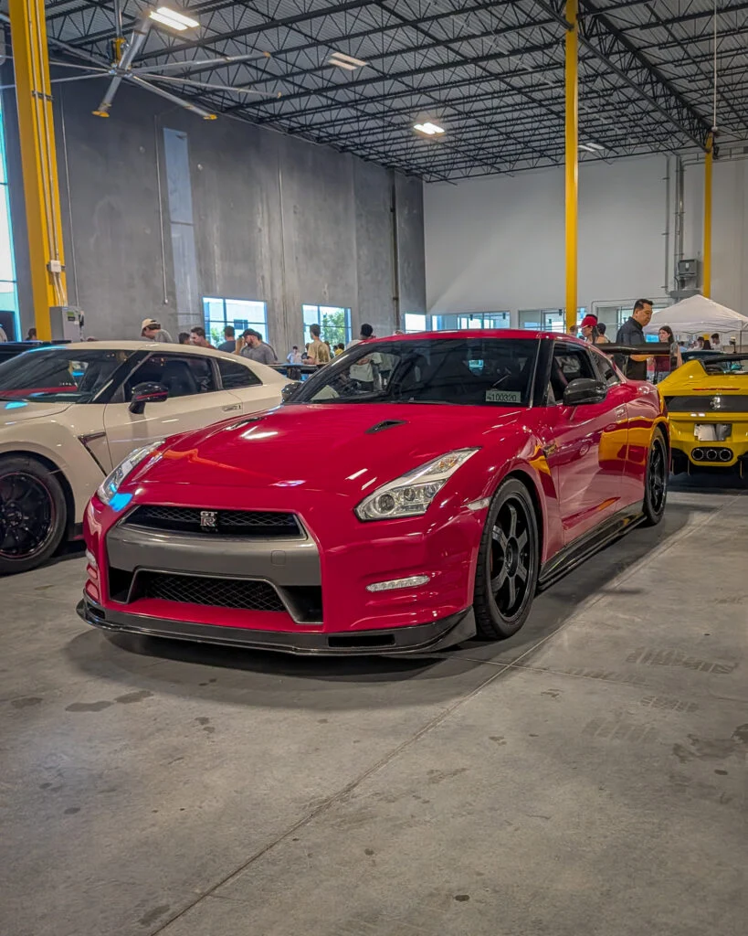 Red Nissan GT-R parked indoors next to a white Nissan GT-R and a yellow Ferrari sports car with people in the background