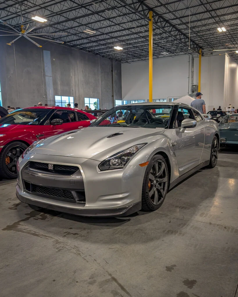 Silver Nissan GT-R parked indoors with red car behind and people in background