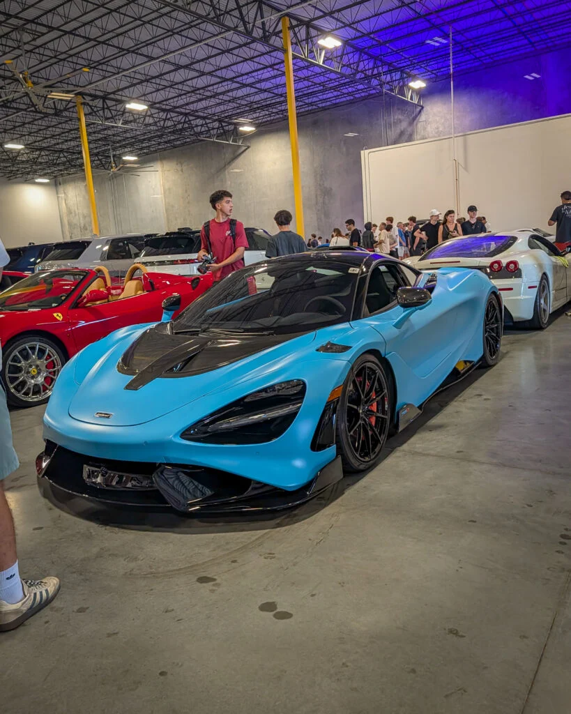 Blue McLaren sports car with black roof parked indoors near red Ferrari convertible and white Ferrari coupe, with people in