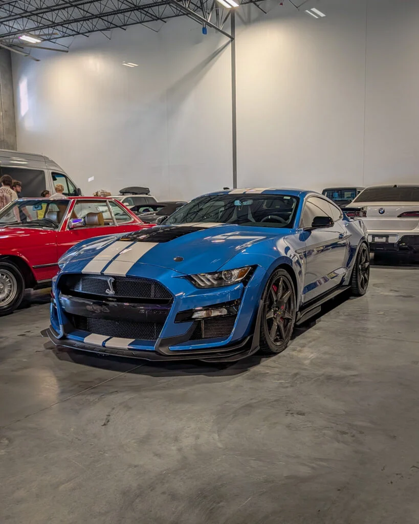 Blue Ford Mustang Shelby GT500 with white racing stripes parked indoors at a car event, with other vehicles and people
