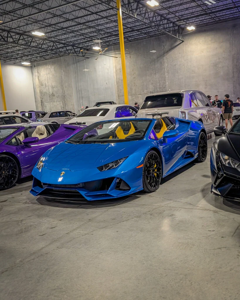 Blue Lamborghini Huracán Spyder with yellow interior parked indoors among several other vehicles and people standing in the
