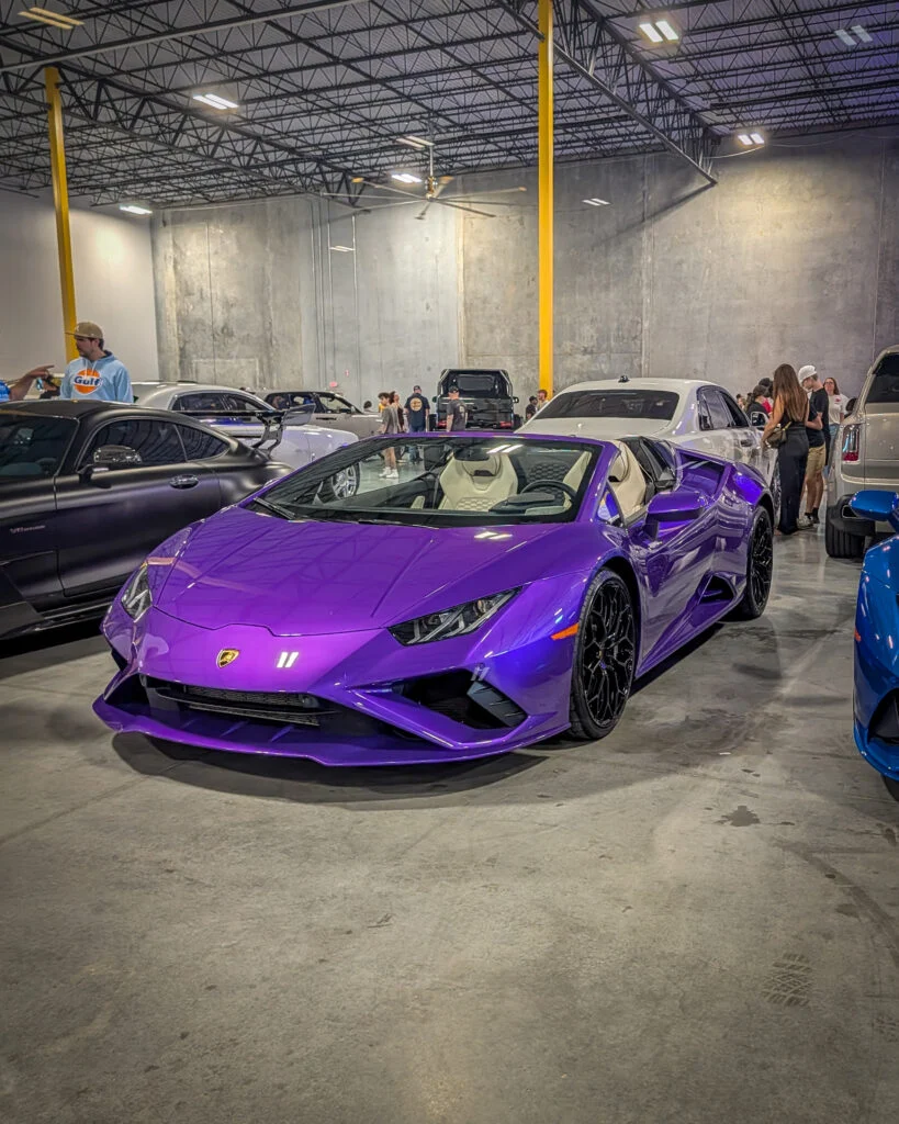 Purple Lamborghini Huracán Spyder parked indoors at a car meet with other vehicles and people in the background