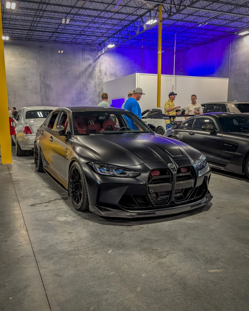 Matte black BMW M3 with red interior inside a warehouse with several people and other cars nearby