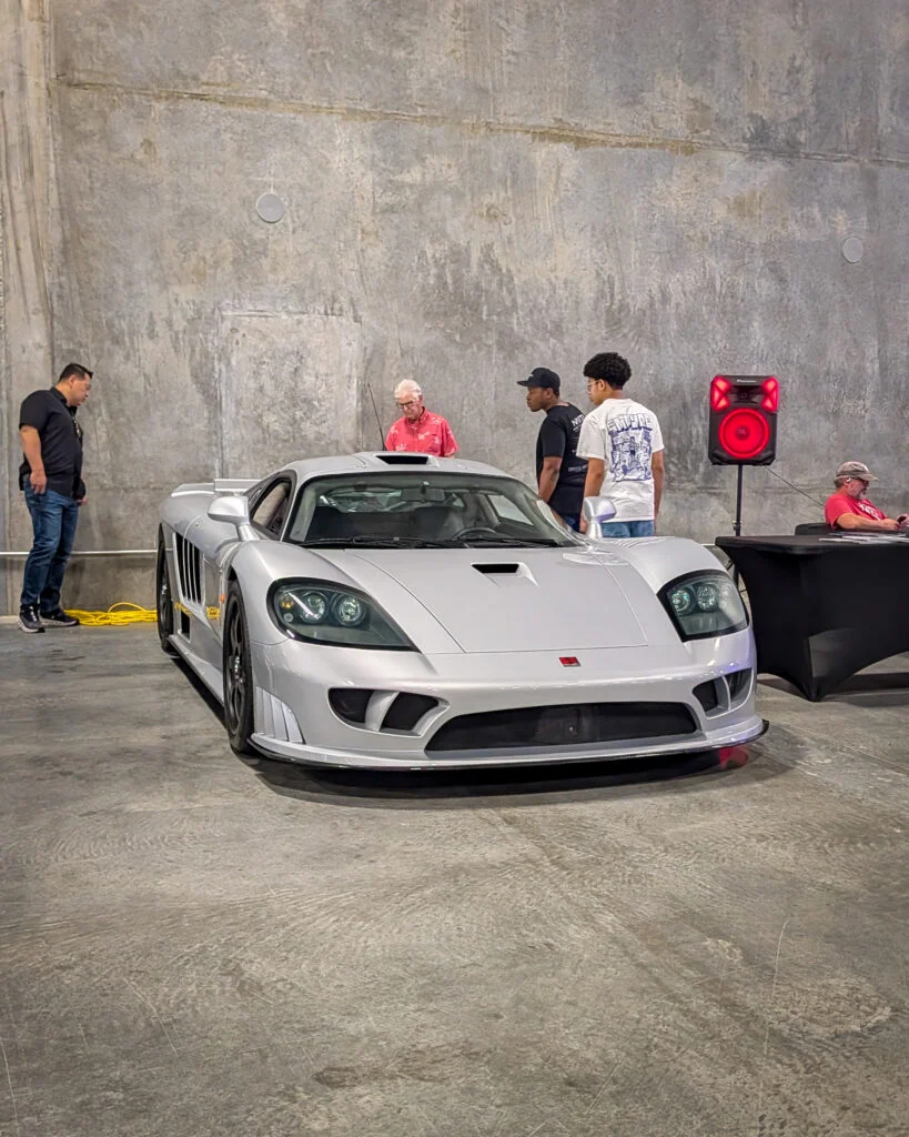 Silver Saleen S7 parked indoors with four men standing near it and one man seated at a table with a red speaker nearby