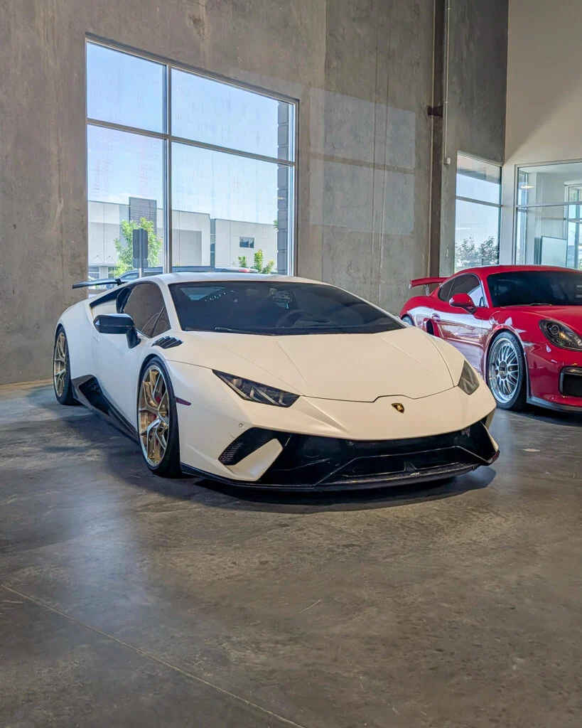 White Lamborghini Huracán and red Porsche Cayman parked indoors by large windows