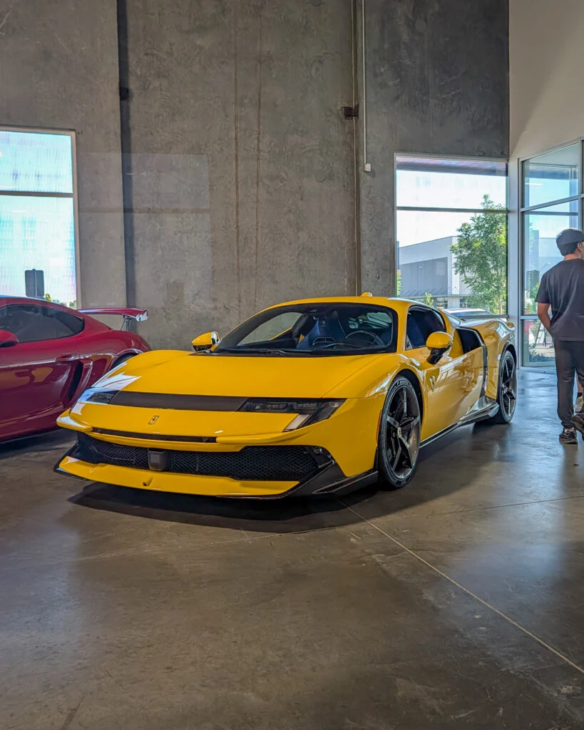Yellow Ferrari sports car front 3/4 parked indoors with a red car beside it and a person standing near glass windows