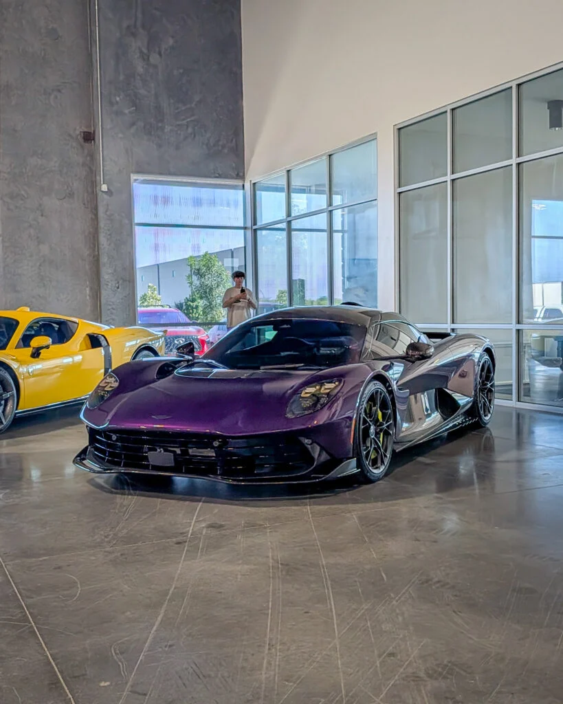 Purple Aston Martin Valhalla and yellow Ford GT parked indoors near glass windows with a person reflected in the glass