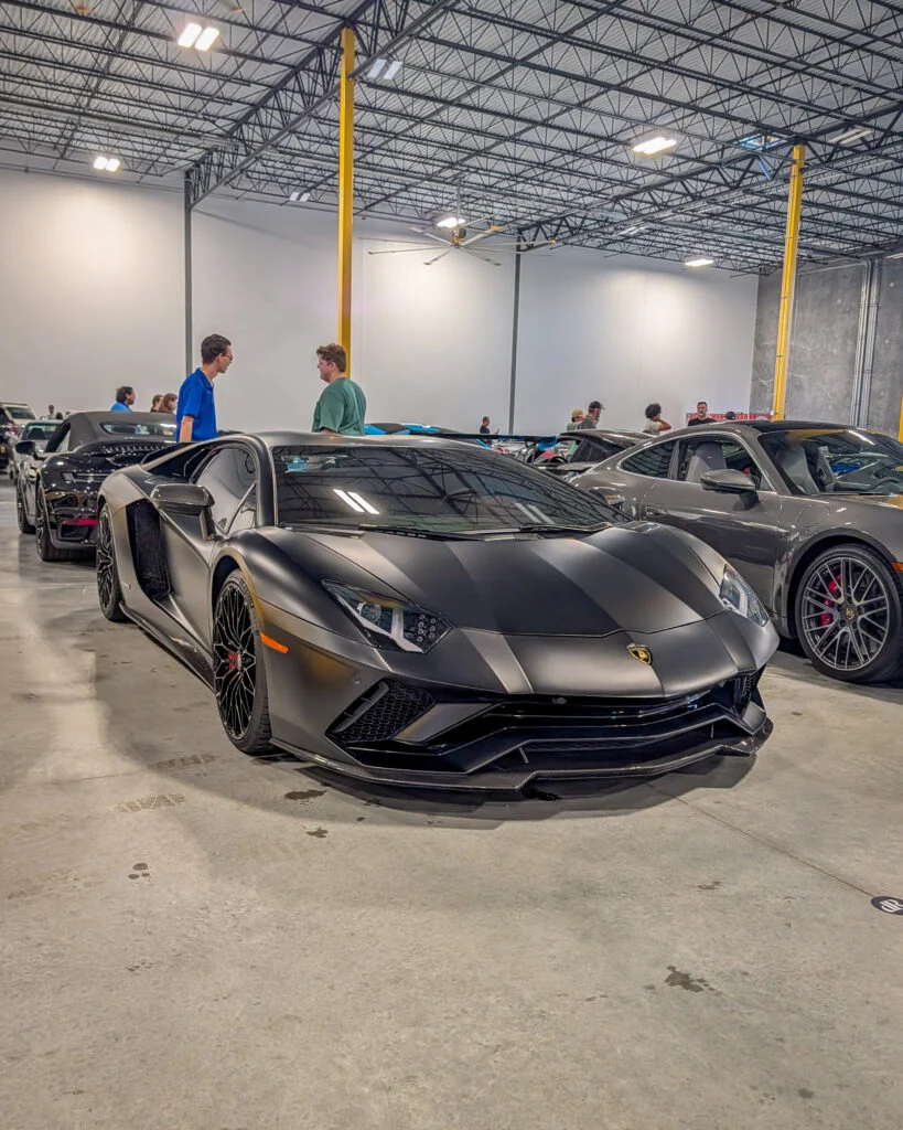 Matte black Lamborghini parked indoors at a car event with people and other vehicles nearby