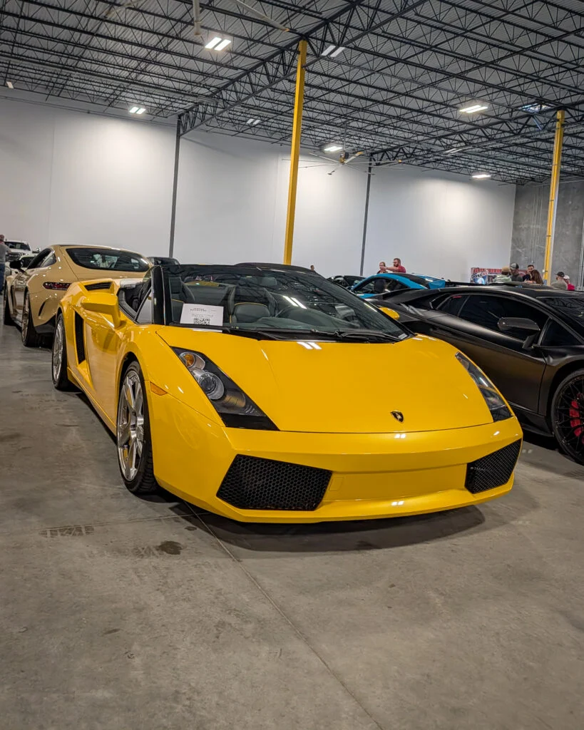 Yellow Lamborghini Gallardo Spyder convertible displayed indoors with other cars and people in the background