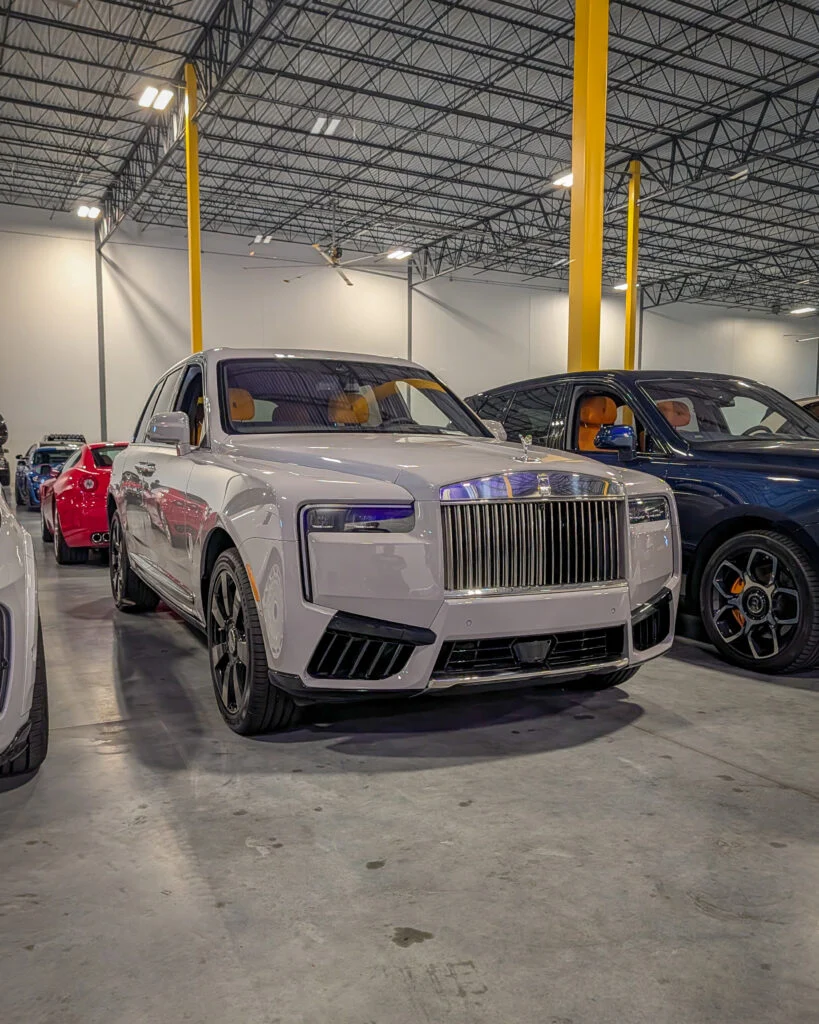White Rolls-Royce Cullinan with tan interior parked indoors next to a blue SUV and red sports car