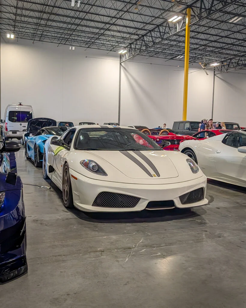 White Ferrari F430 with black racing stripes parked indoors among other vehicles and people.