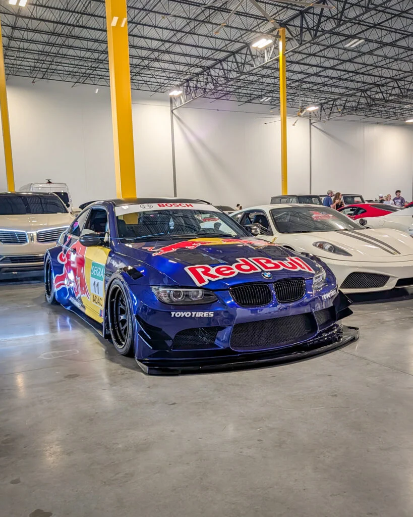 Blue Red Bull BMW race car and white Ferrari coupe indoors at a car event with people in background