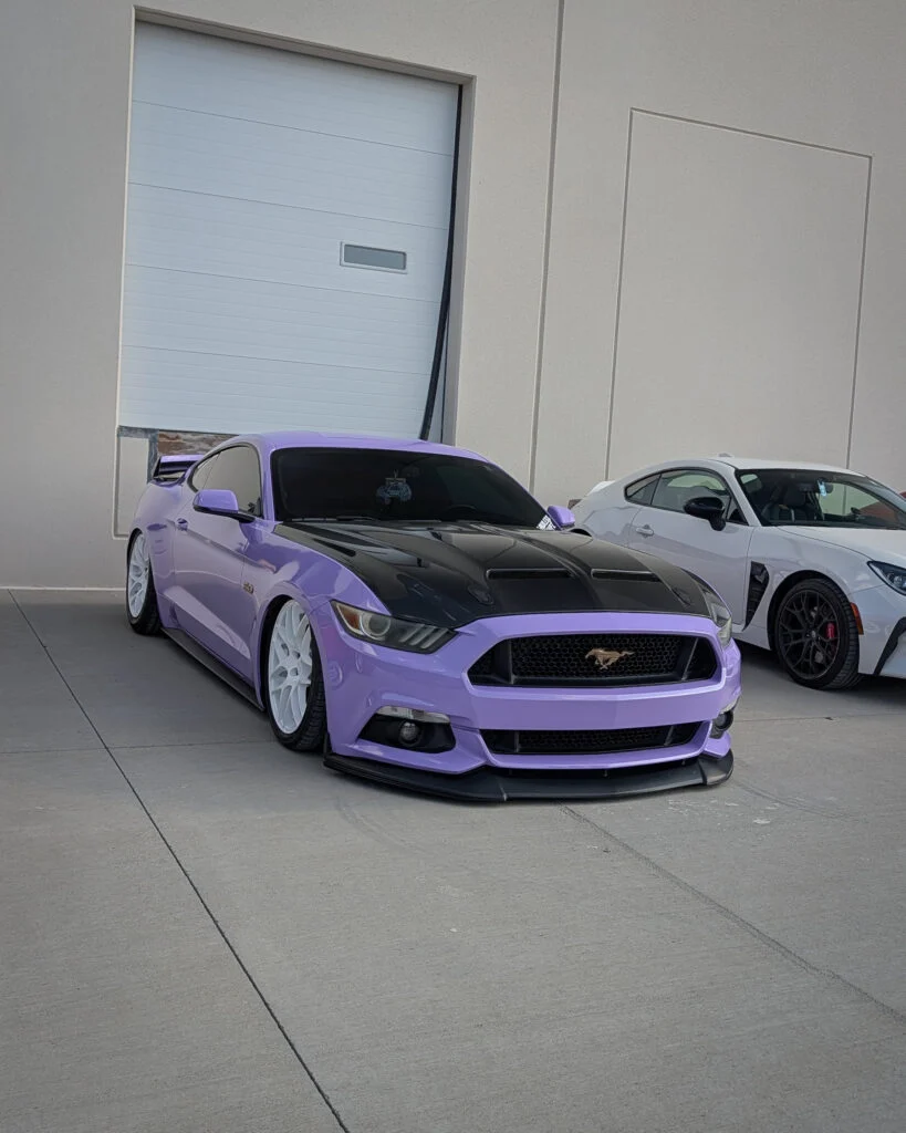 Lavender Ford Mustang with black hood and white wheels parked next to a white coupe near a beige wall with a closed garage