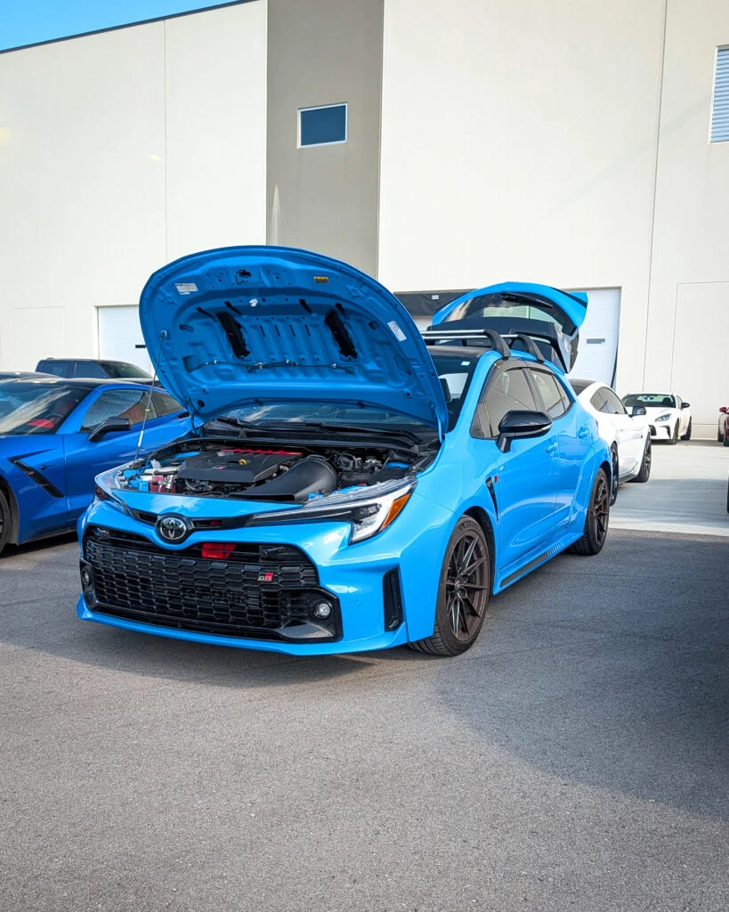 Blue Toyota GR Corolla with hood open, white coupe behind it, and blue coupe parked next to it near a beige building.