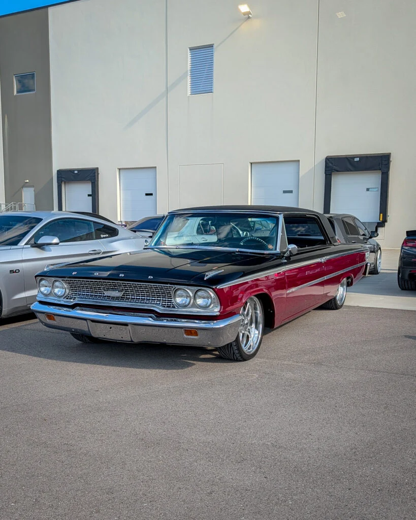 Two-tone black and red Ford Galaxie 500 parked next to silver coupe in a lot outside a beige building with loading doors.
