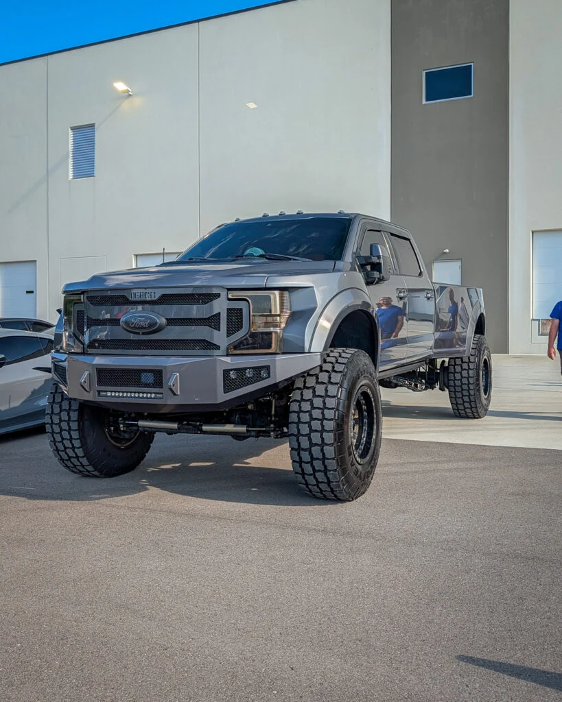 Gray Ford F-150 pickup truck with large off-road tires parked in front of a beige building under blue sky