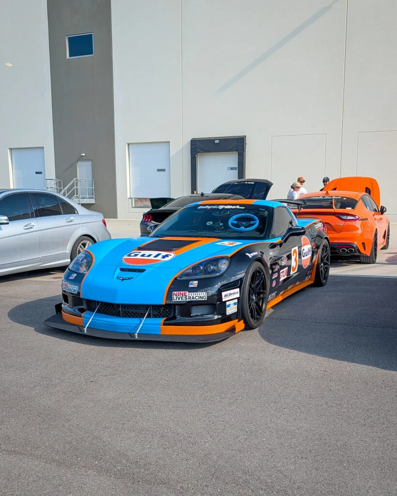 Blue and orange Gulf-liveried Chevrolet Corvette race car and orange Porsche sedan parked outdoors, with people nearby and