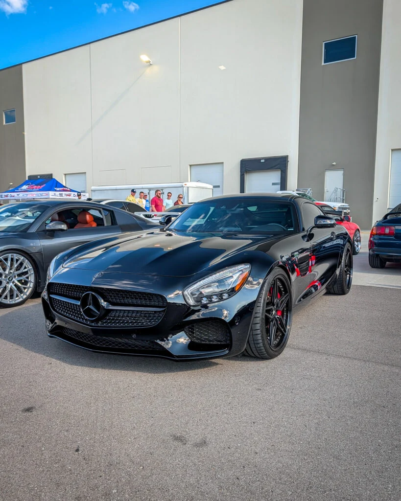 Black Mercedes-Benz AMG GT parked at a car event with other vehicles and people nearby against a warehouse backdrop.
