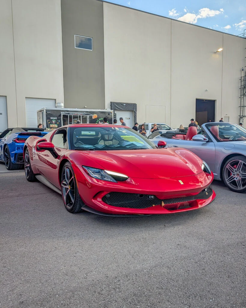 Red Ferrari 296 GTB parked next to a silver convertible and blue coupe against a warehouse backdrop with people nearby.
